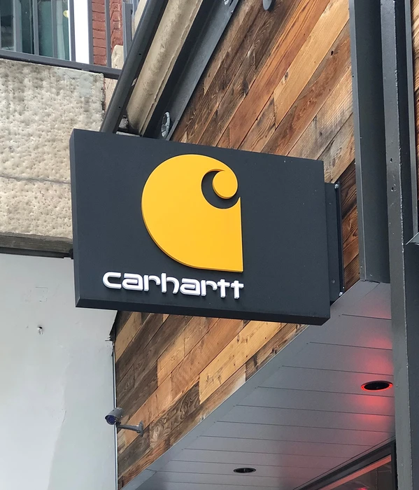 Enseigne commerciale Carhartt fixée sur façade urbaine avec logo jaune distinctif en forme de "C" stylisé sur fond noir