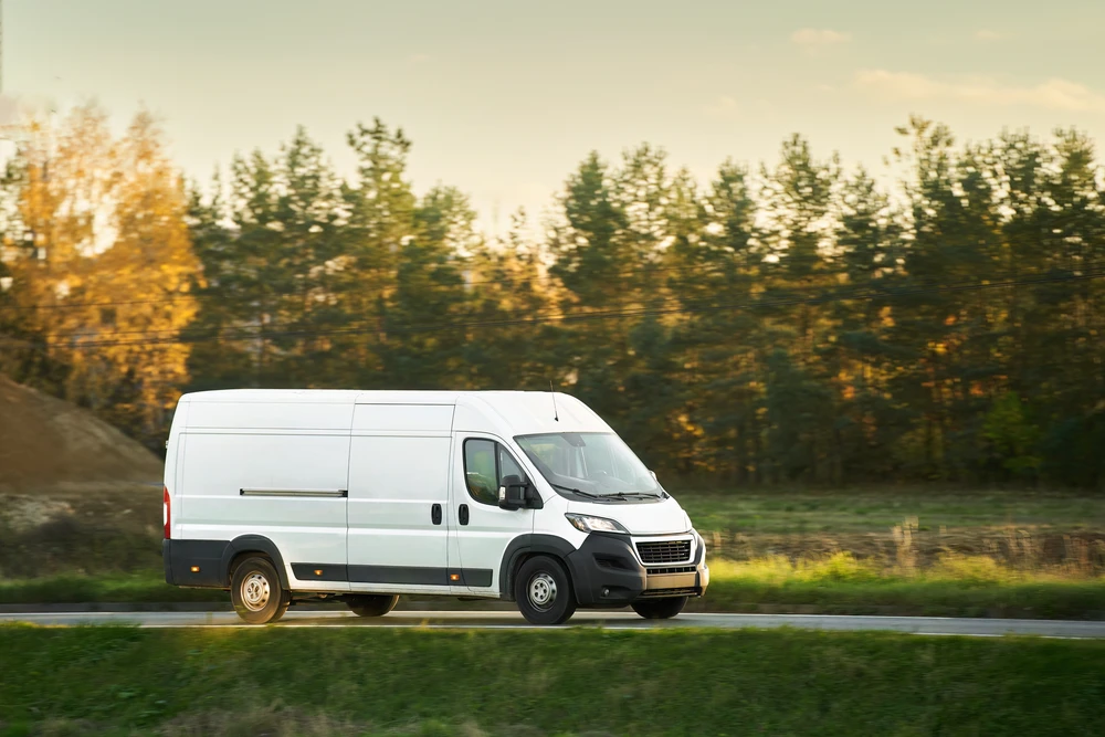 Fourgon utilitaire blanc circulant sur route de campagne au coucher du soleil, environnement rural avec arbres et végétation en arrière-plan